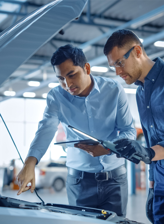 A technician and an advisor looking over a car engine with tablets.