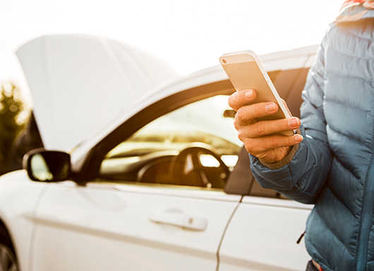 Person standing next to car with hood up, looking at smartphone.