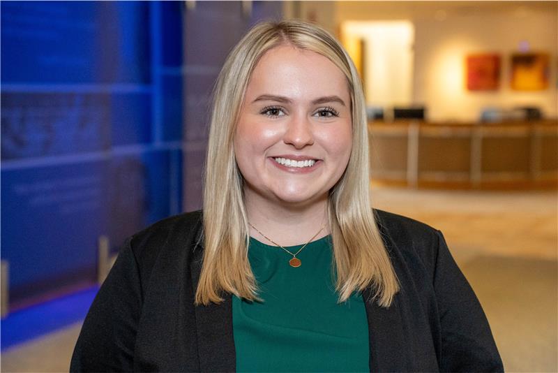 Headshot of a young woman with blonde hair, wearing a black blazer and green top.