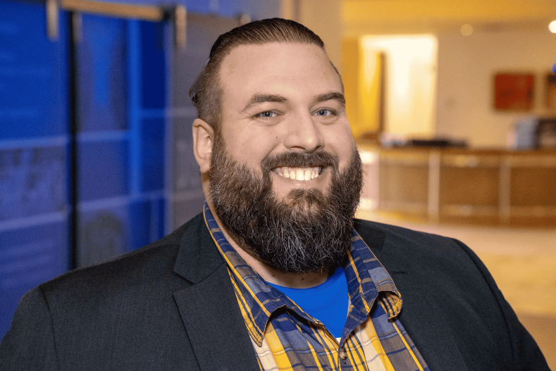Headshot of a man with a brown beard in a blazer with a blue and yellow shirt.